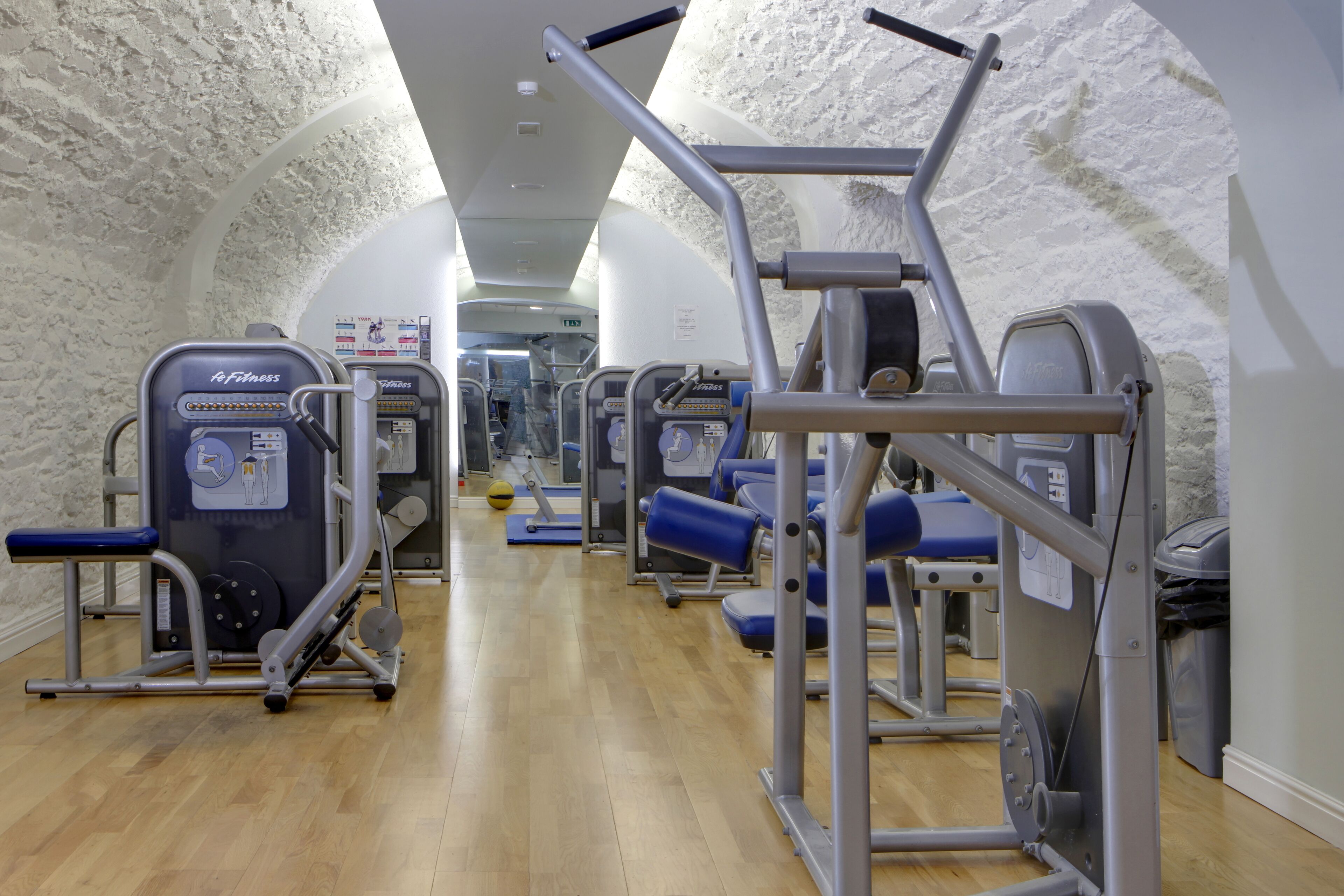 A gym with various exercise machines, a wooden floor, and white walls.