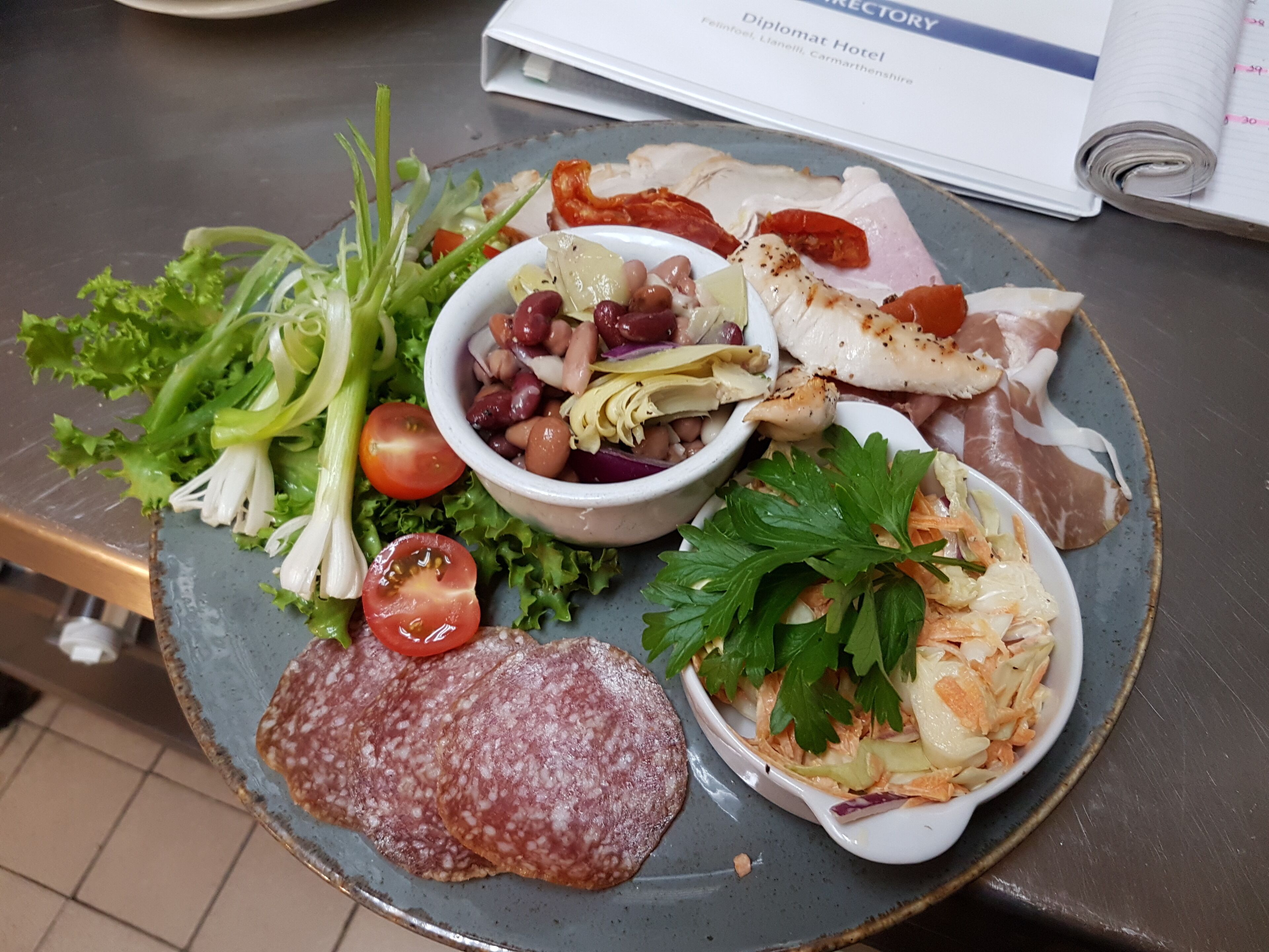 A plate of food with various items including sliced salami, a salad, and a bowl of beans.
