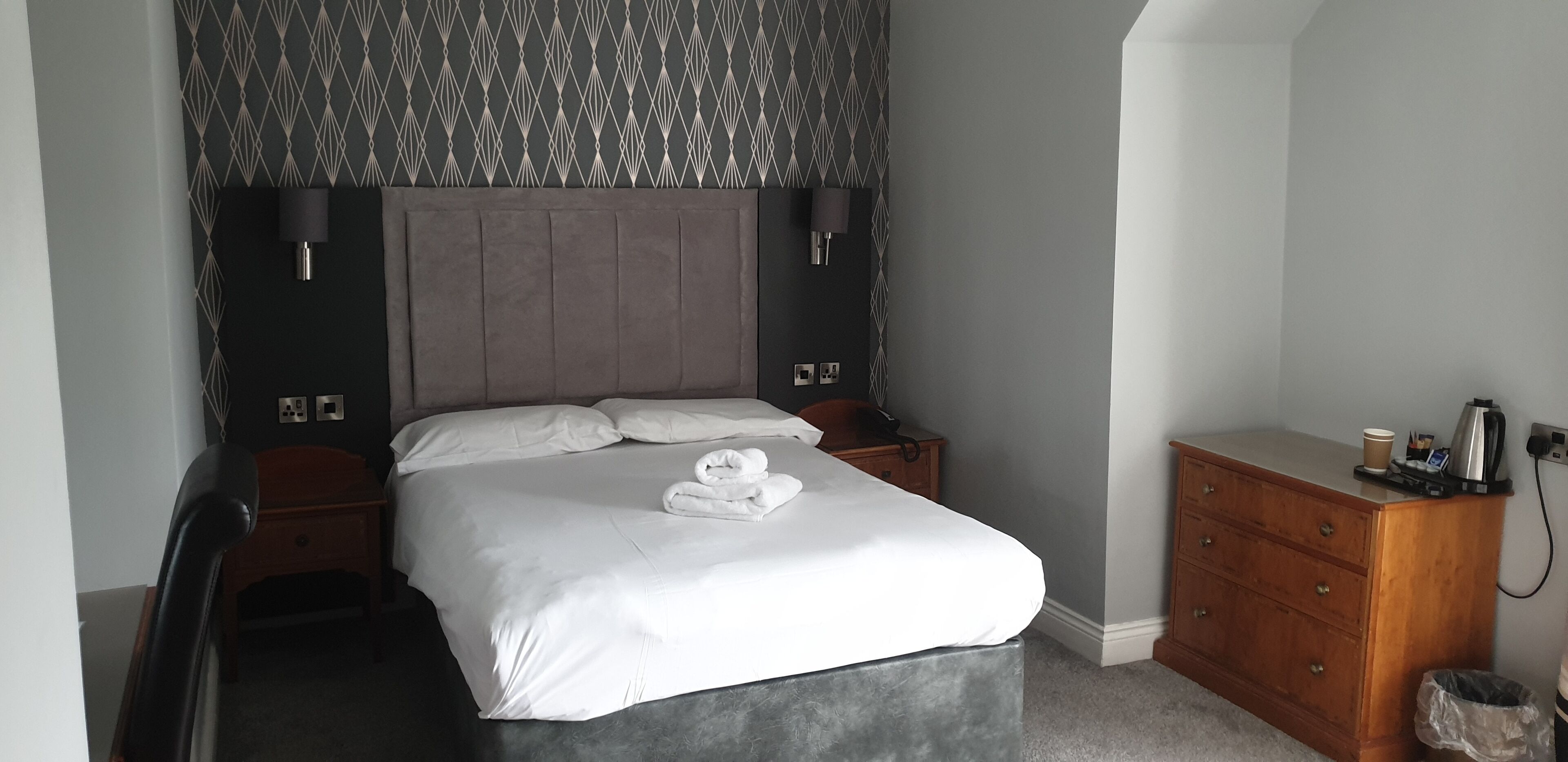 A neatly made bed with white linens and a folded towel, a wooden nightstand with a lamp, a dresser with a cup and a kettle, and a patterned wallpaper.