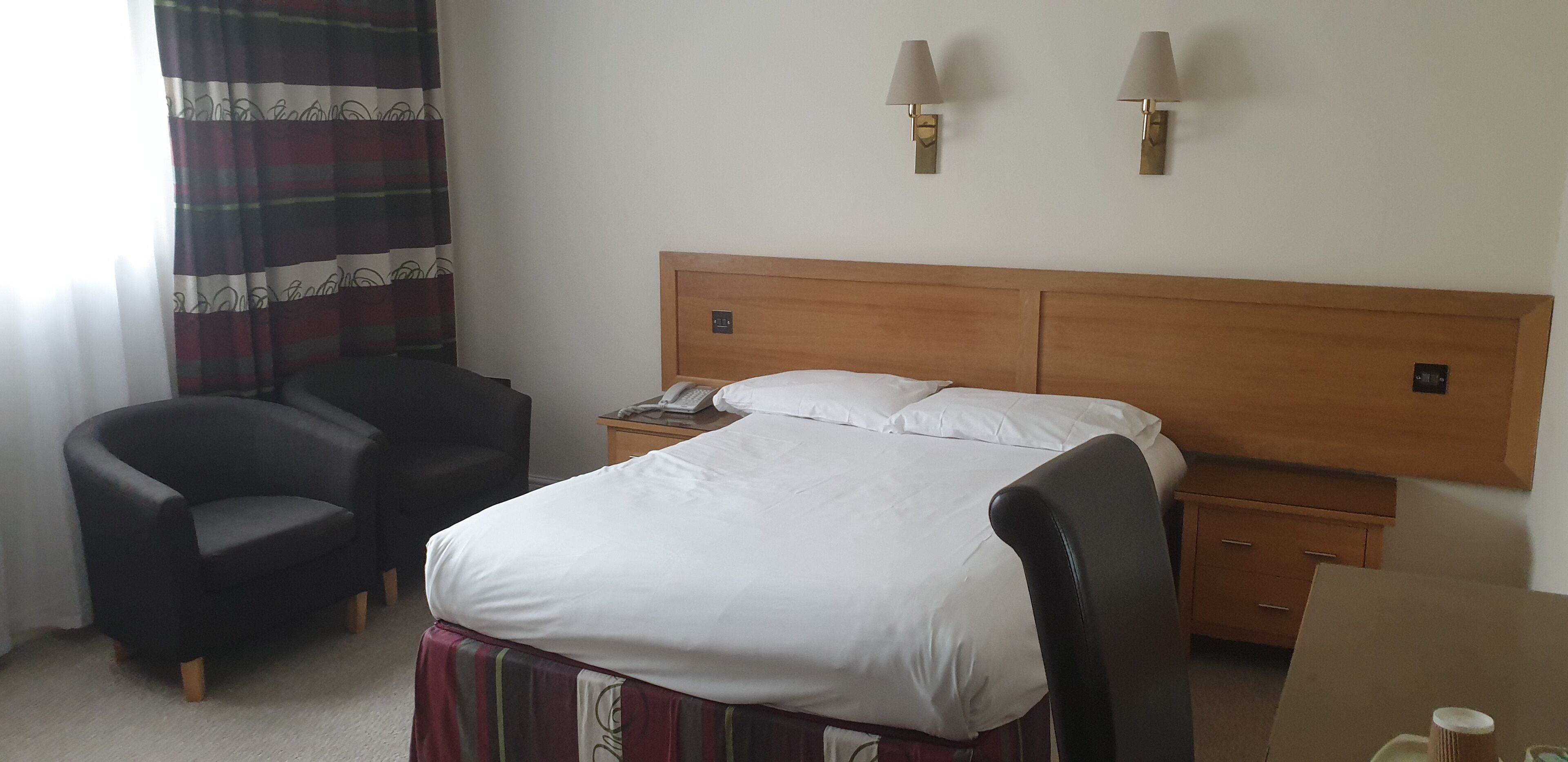 A hotel room with a bed, a chair, a nightstand with a phone, and two wall-mounted lamps.