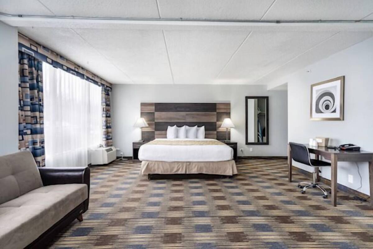 A hotel room with a bed, a desk, a chair, a sofa, and a large window with curtains.