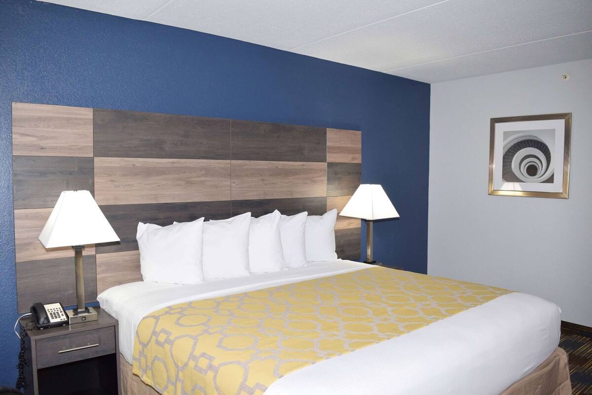 A hotel room with a bed, two bedside lamps, a nightstand with a phone, and a wall-mounted picture.