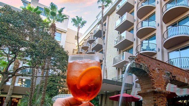 A hand holding a glass with a drink, a fountain, and a building with balconies. - Hotel Lucerna Tijuana (Tijuana)