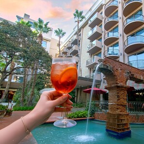 A hand holding a glass with a drink, a fountain, and a building with balconies.