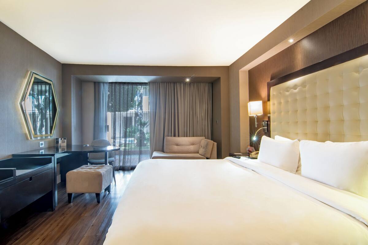 Ein Hotelzimmer mit einem großen Bett, einem Schreibtisch, einem Sessel und Blick ins Freie.