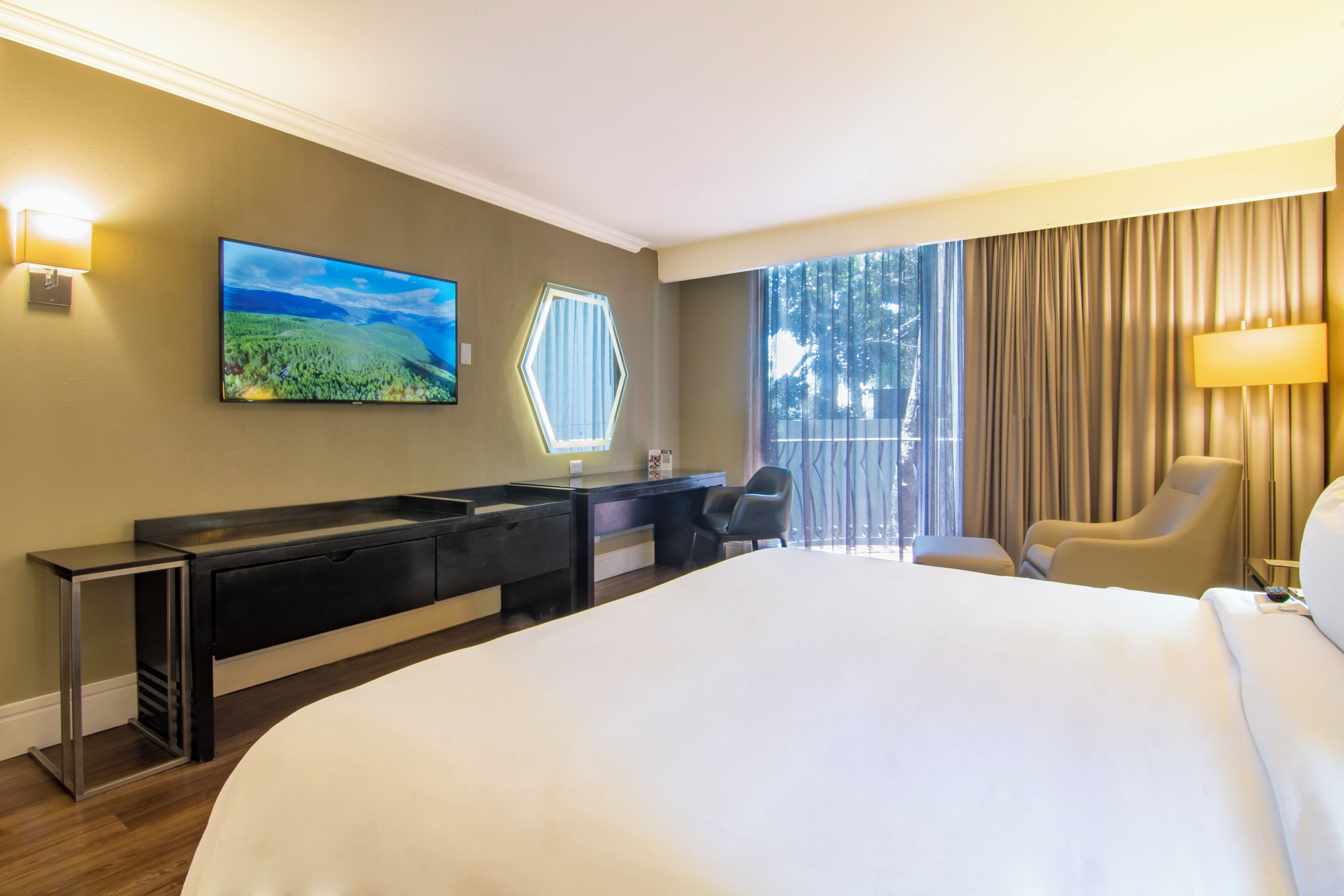 A hotel room with a large bed, a desk with a chair, a television, and a window with curtains.