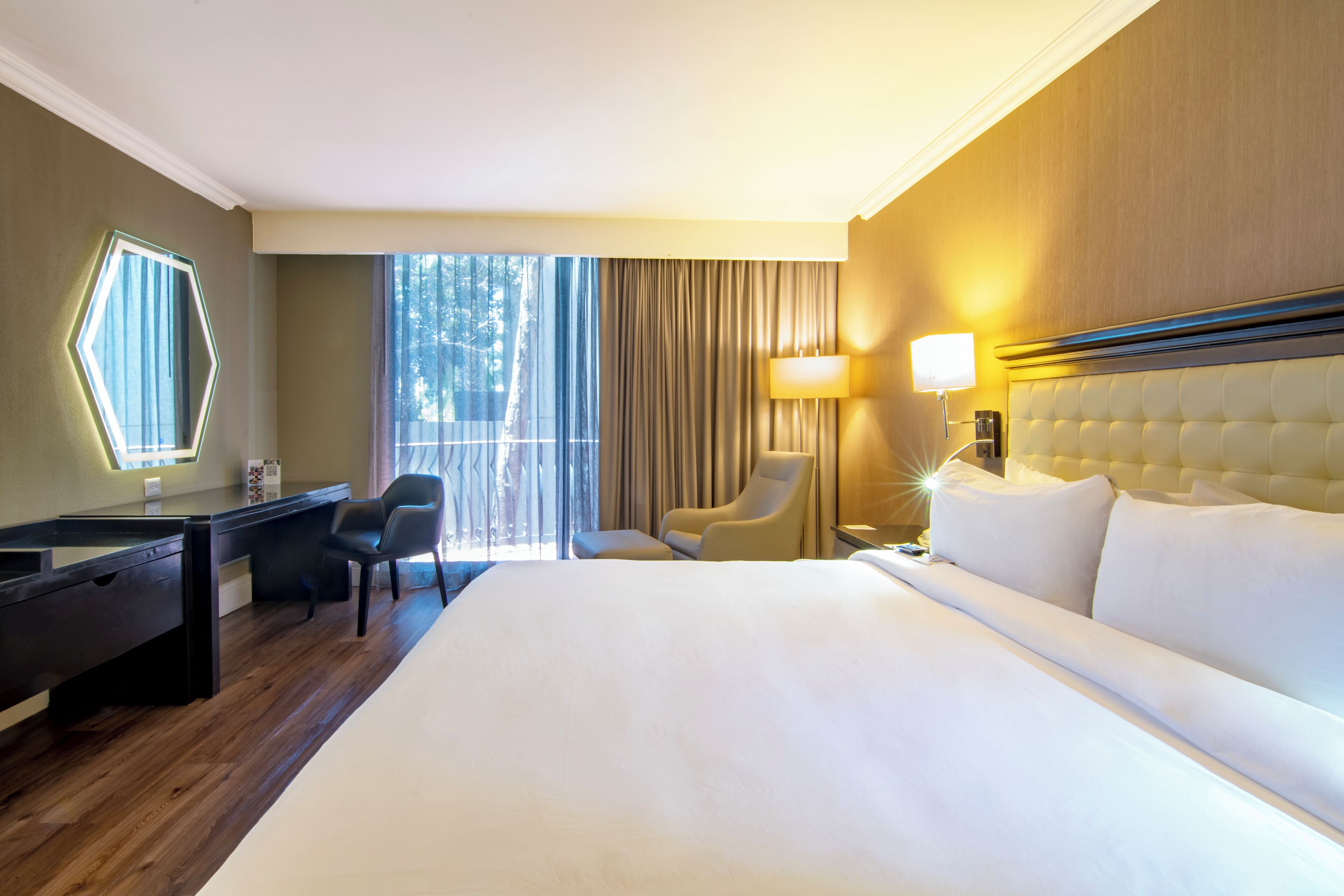 Habitación de hotel con una cama grande, un escritorio con silla, una lámpara y vistas al exterior.