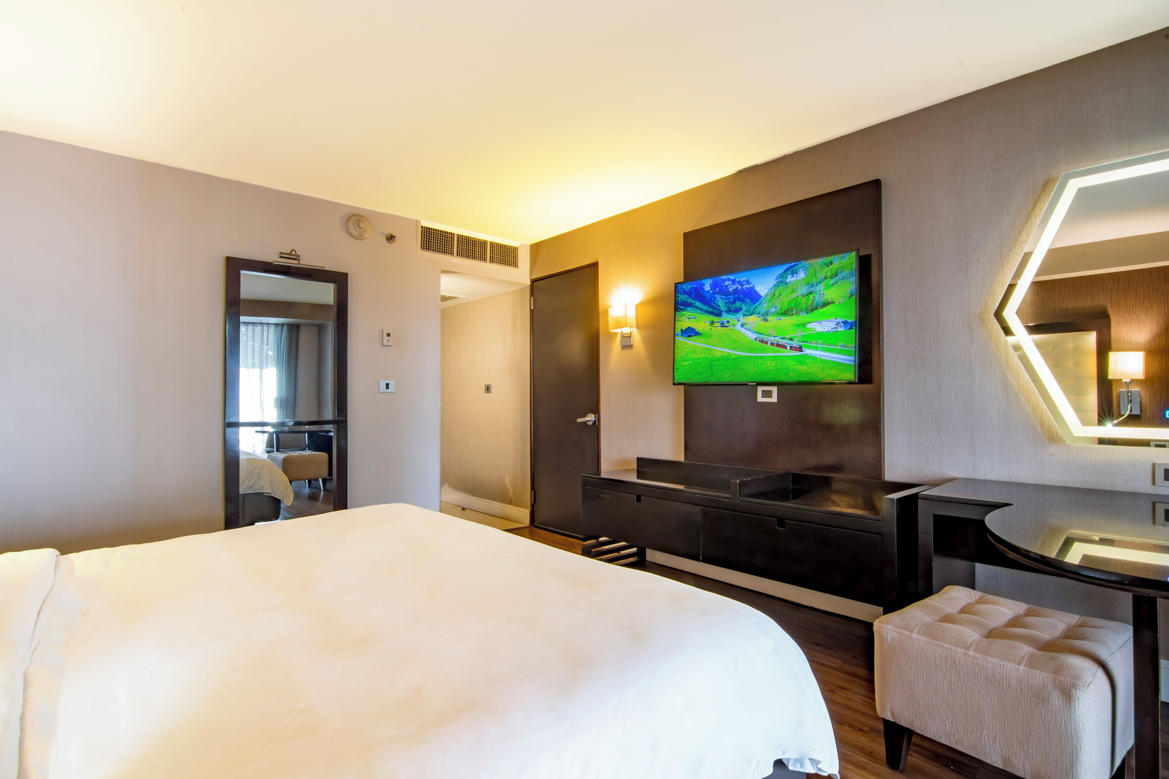 A modern hotel room with a large bed, a TV mounted on the wall, a desk with a chair, and a mirror with a geometric light fixture.