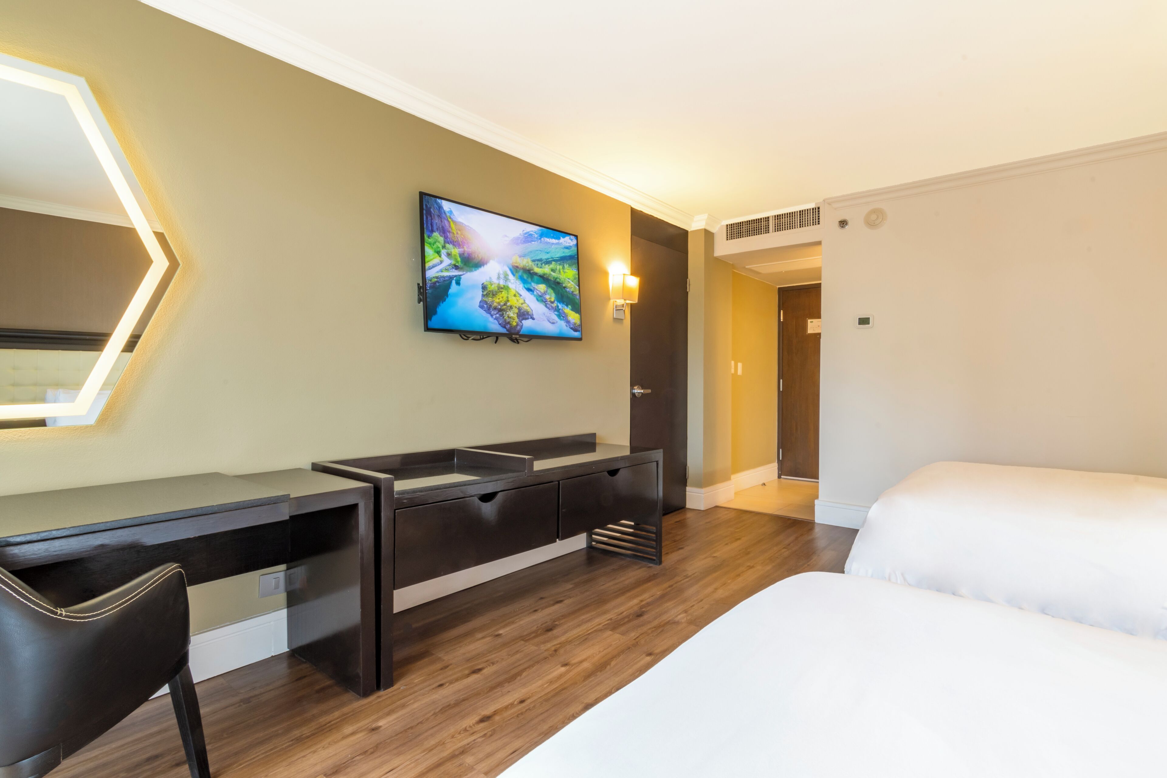 Una habitación de hotel moderna con suelo de madera, una cama grande, un escritorio con una silla, un televisor montado en la pared y una puerta que da a otra habitación.