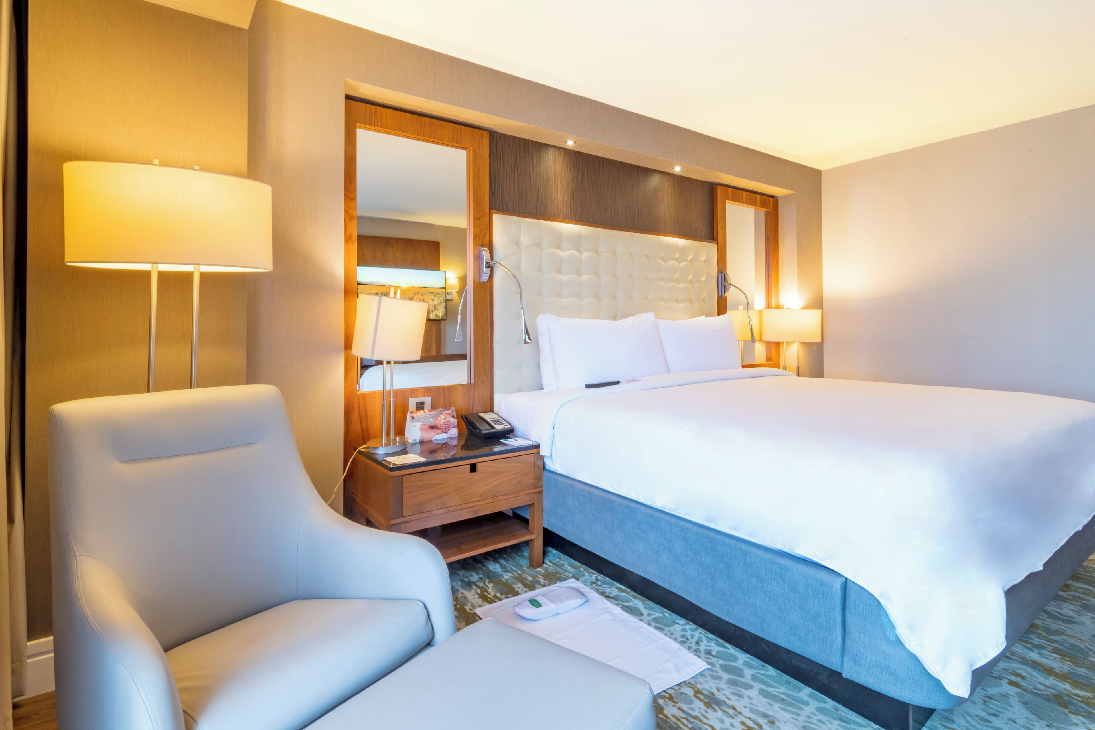 A hotel room with a large bed, a chair, a nightstand with a lamp, and a mirror.