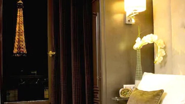 A hotel room with a bed, a nightstand, a lamp, and a view of the Eiffel Tower. - Hôtel Eiffel Trocadéro (Paris)