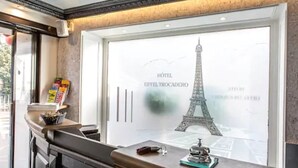 A hotel reception area with a large window displaying the Eiffel Tower, a reception desk, and a small shelf with items. - Hôtel Eiffel Trocadéro (Paris)