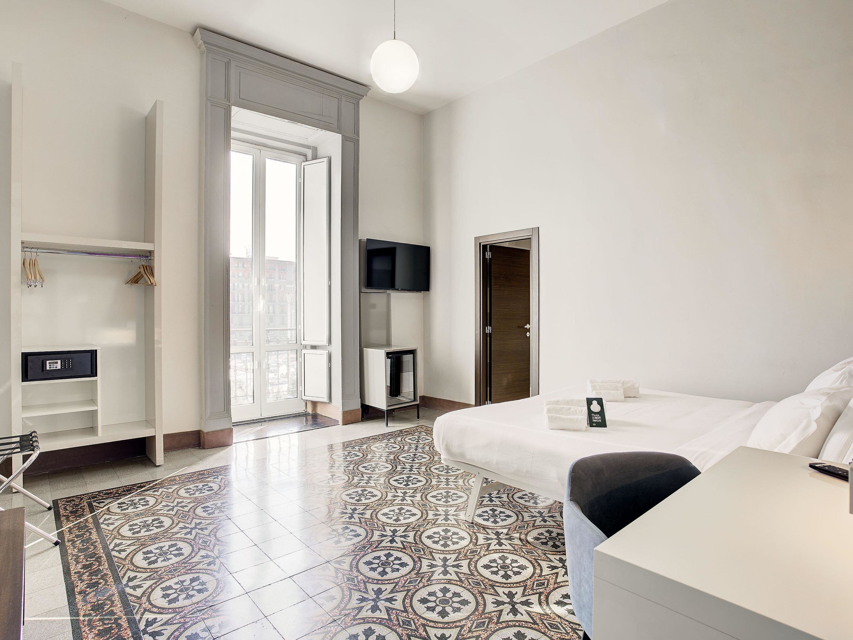 A hotel room with a bed, a desk, a TV, a wardrobe, and a patterned floor.