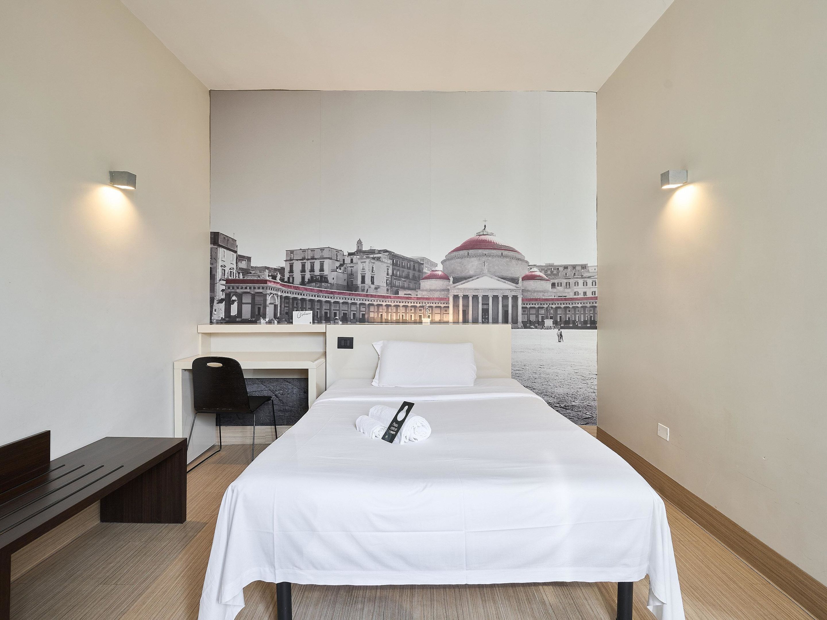 A hotel room with a bed, a bench, a chair, and a large cityscape mural on the wall.