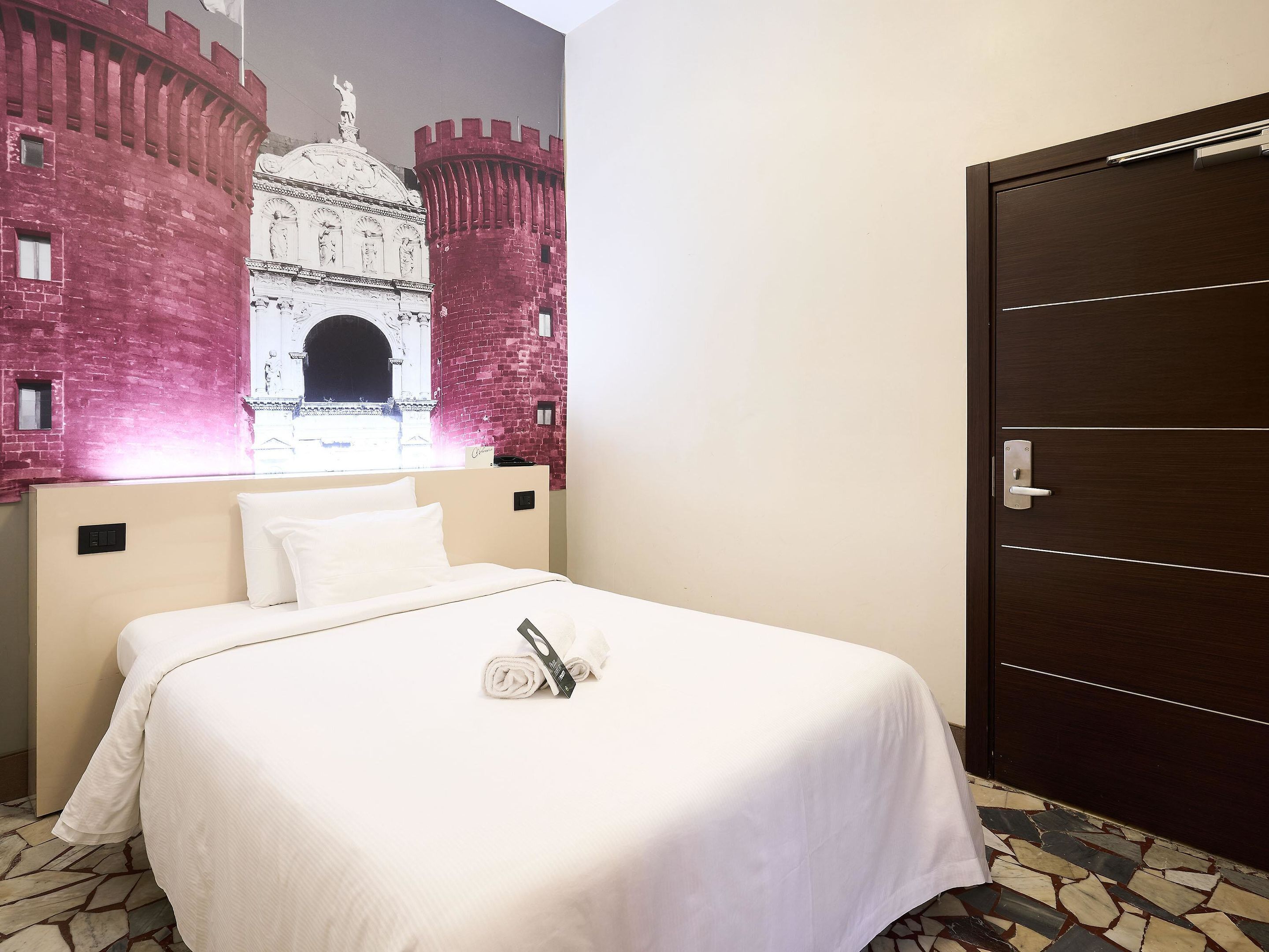 A hotel room with a bed, a large mural of a castle, a wooden door, and a tiled floor.