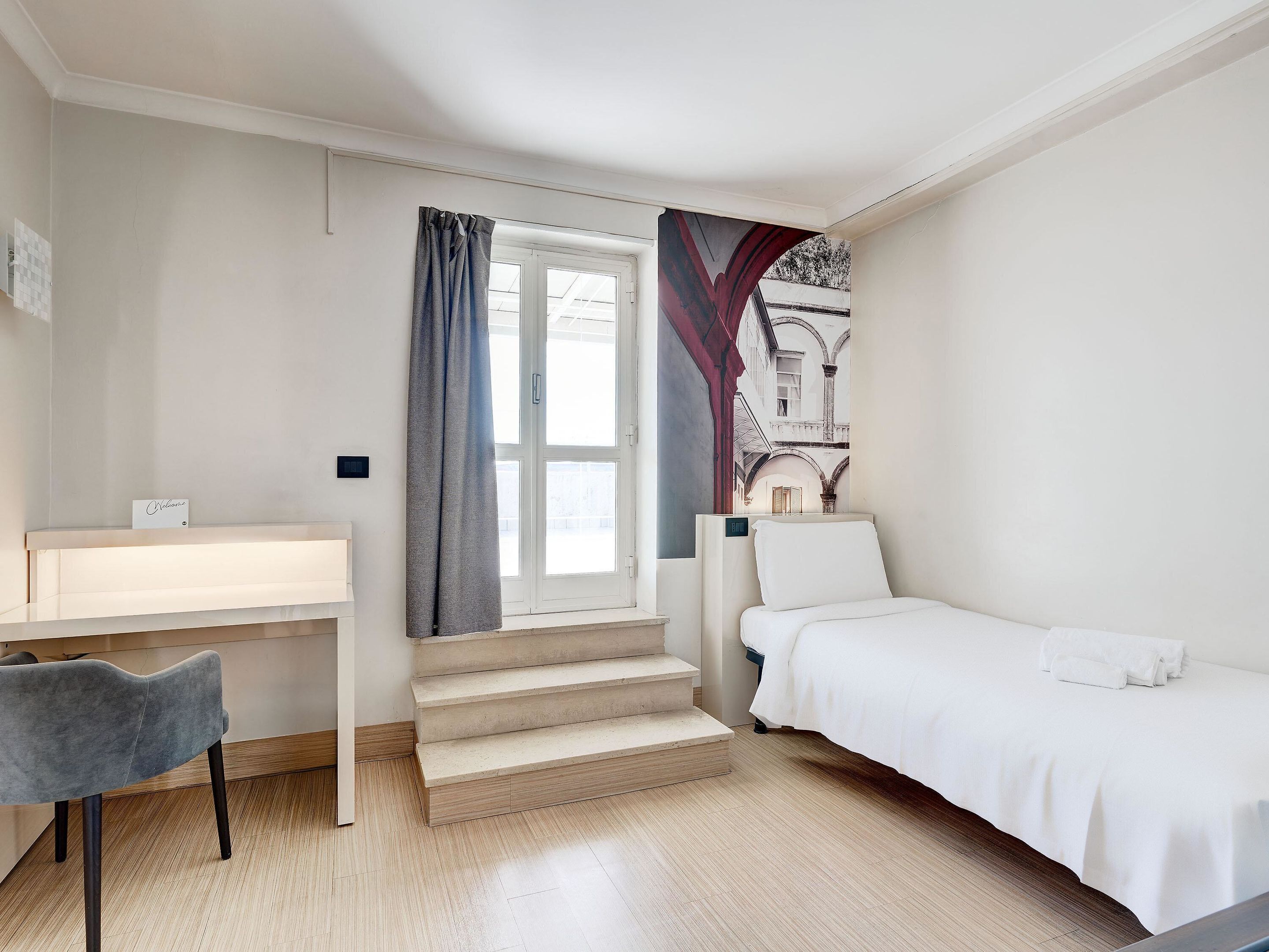 Habitación de hotel con cama, escritorio y una silla. Tiene una ventana con cortinas, un cuadro en la pared y una escalera.