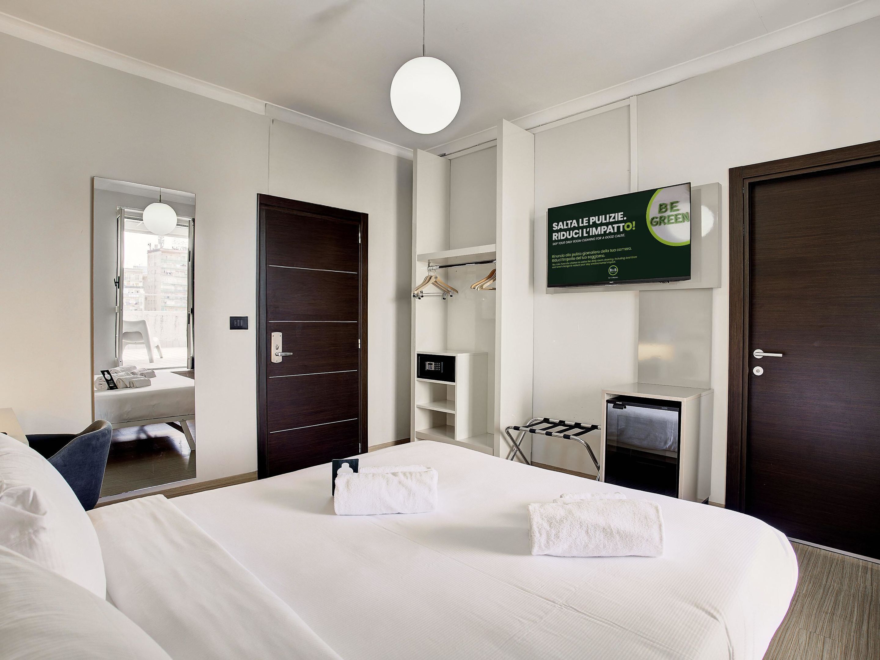 A hotel room with a bed, a mirror, a wardrobe, a towel rack, and a sign promoting eco-friendly practices.