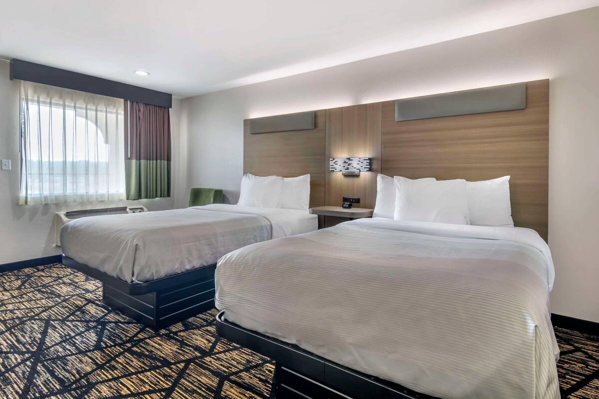 A hotel room with two beds, a large window with curtains, a flat-screen TV, and a patterned carpet.