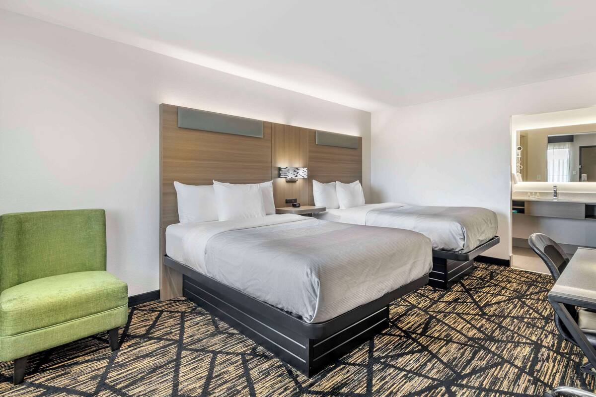 A hotel room with two beds, a green armchair, a desk, and a mirror.
