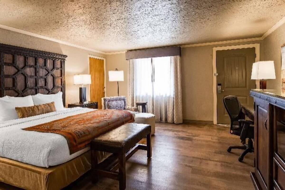 A hotel room with a large bed, a wooden headboard, a desk with a chair, a lamp, and a painting on the wall.