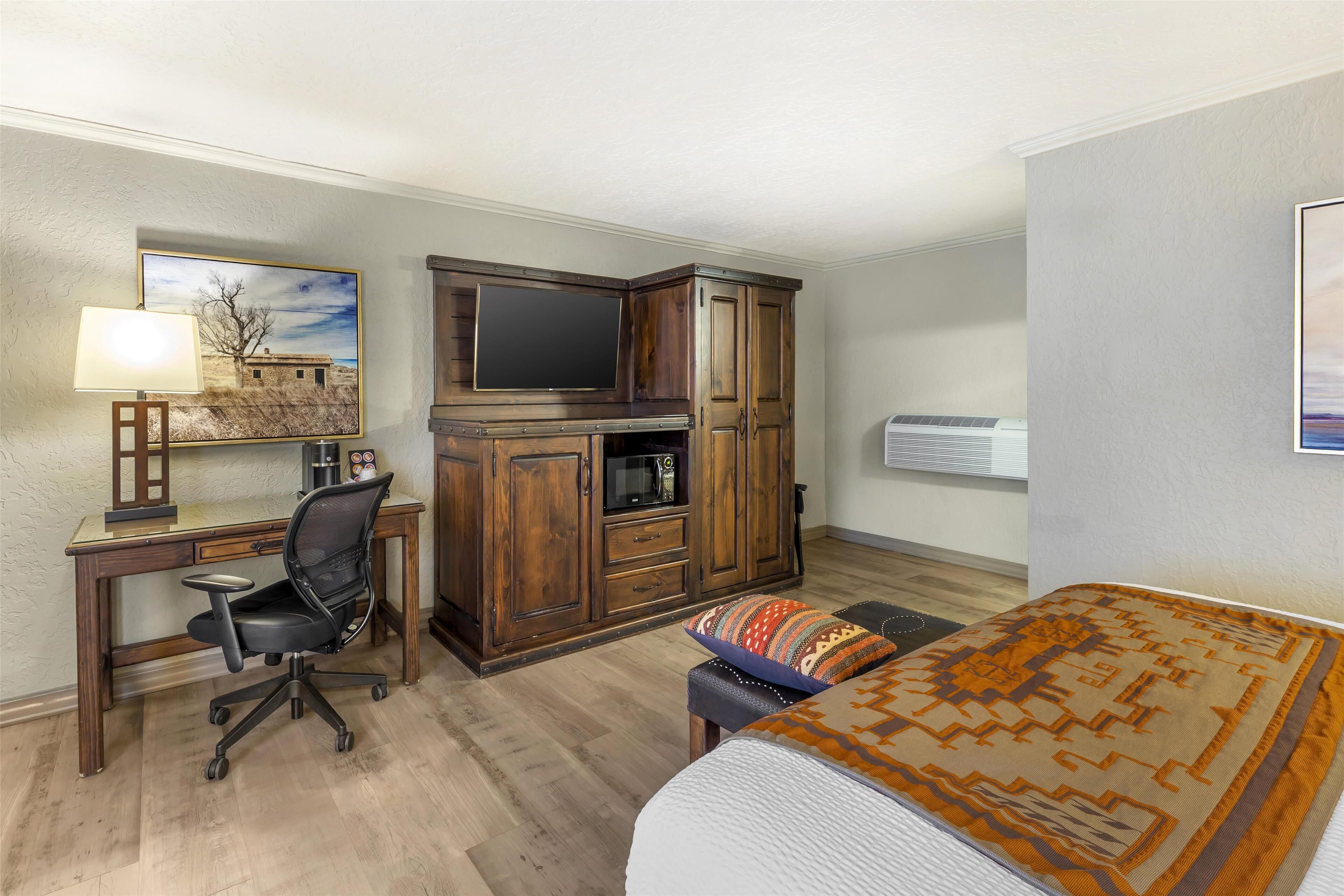 A hotel room with a wooden desk, a black office chair, a bed with patterned bedding, a television, and a large window.