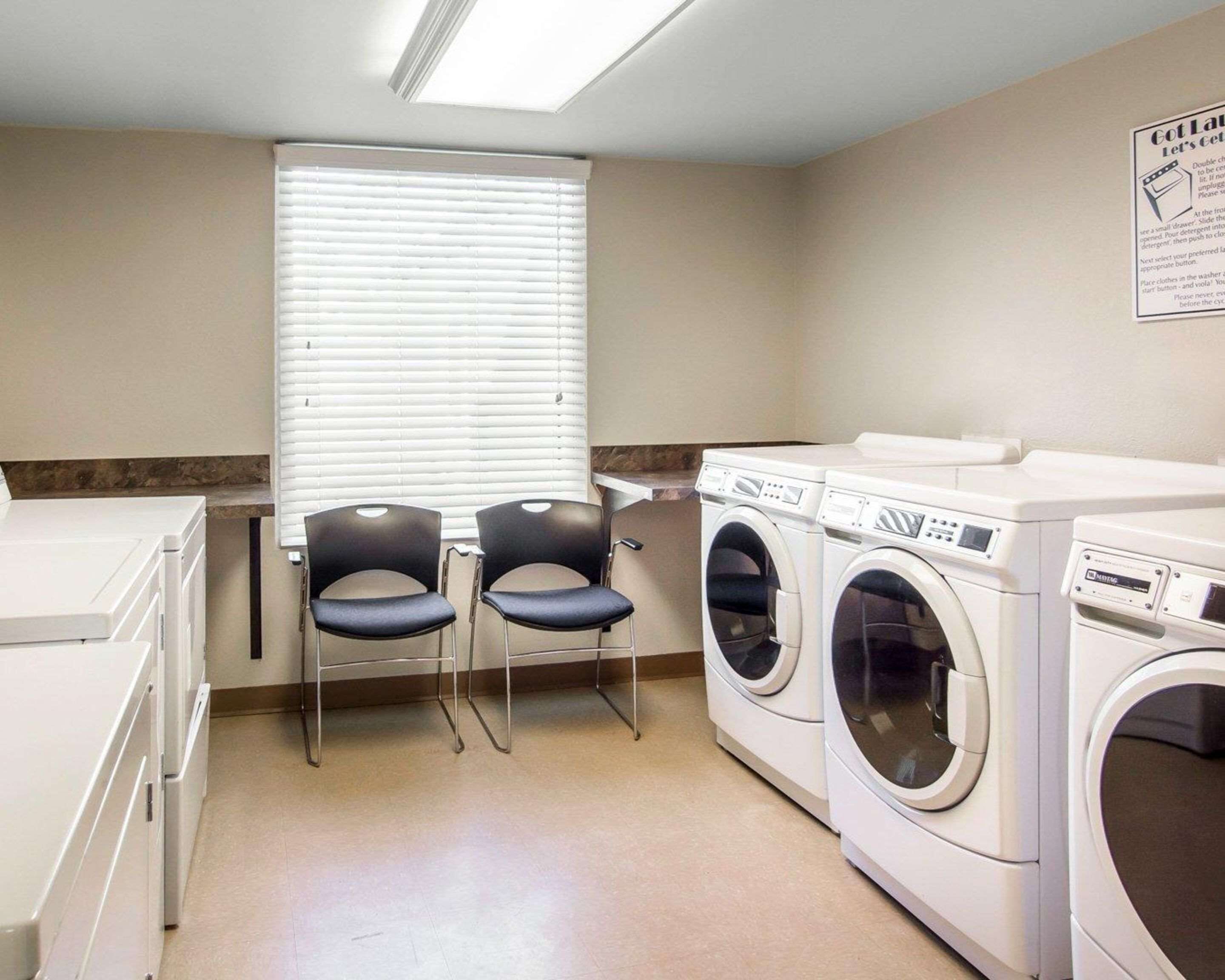 A laundry room with two chairs, a window with blinds, and two front-loading washing machines.