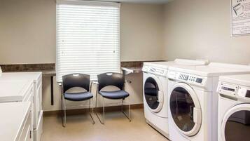 A laundry room with two chairs, a window with blinds, and two front-loading washing machines.