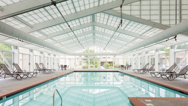 An indoor swimming pool with clear glass walls, multiple lounge chairs, and a high ceiling with skylights. - Bluegreen Vacations Patrick Henry Square, an Ascend Collection Resort (Williamsburg)