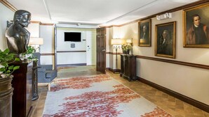 A hotel hallway with a patterned carpet, a bust sculpture, framed portraits, and a television on the wall. - Bluegreen Vacations Patrick Henry Square, an Ascend Collection Resort (Williamsburg)