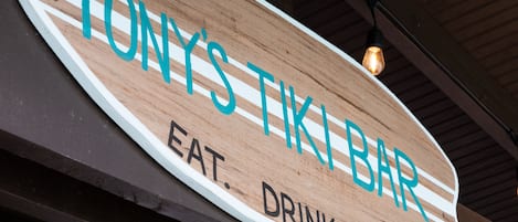 Uma placa de madeira com o texto 'TONY'S TIKI BAR' e 'COMER. BEBER. FÉRIAS'.