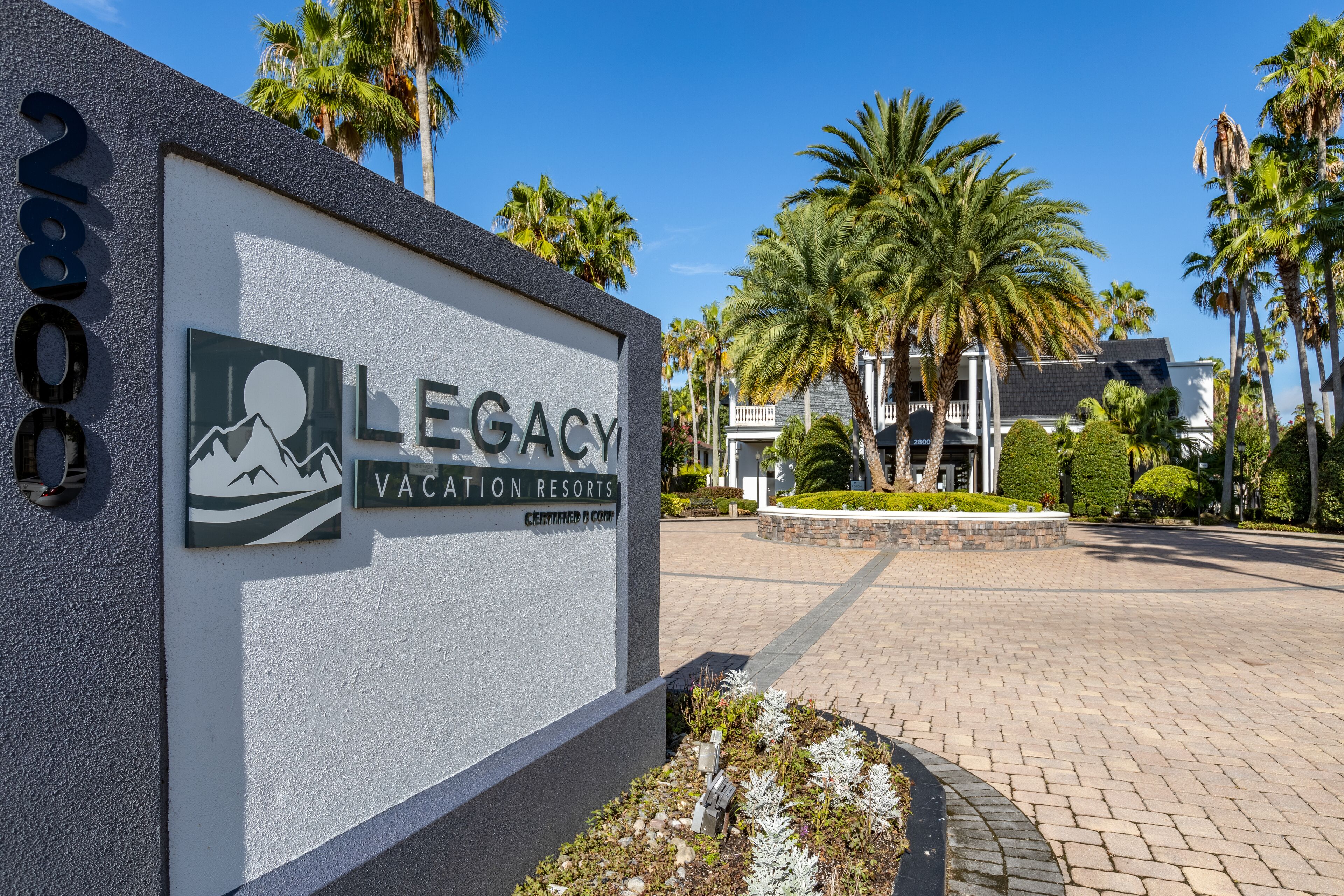 A sign for Legacy Vacation Resorts with a mountain graphic, palm trees, and a paved driveway.