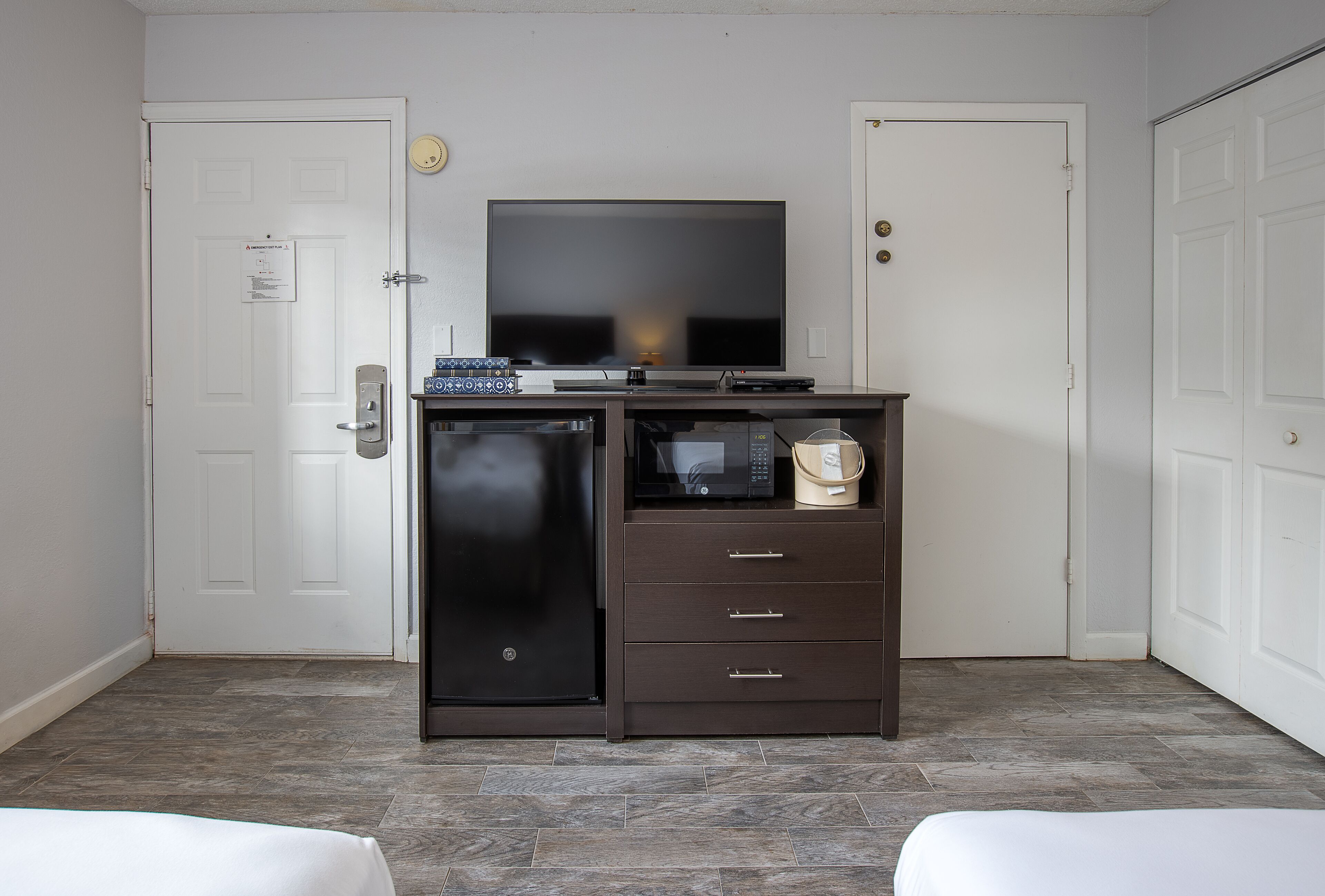 Habitación compacta con televisor sobre un mueble, microondas y refrigerador pequeño. Se observan dos puertas blancas y una pared blanca con detector de humo.
