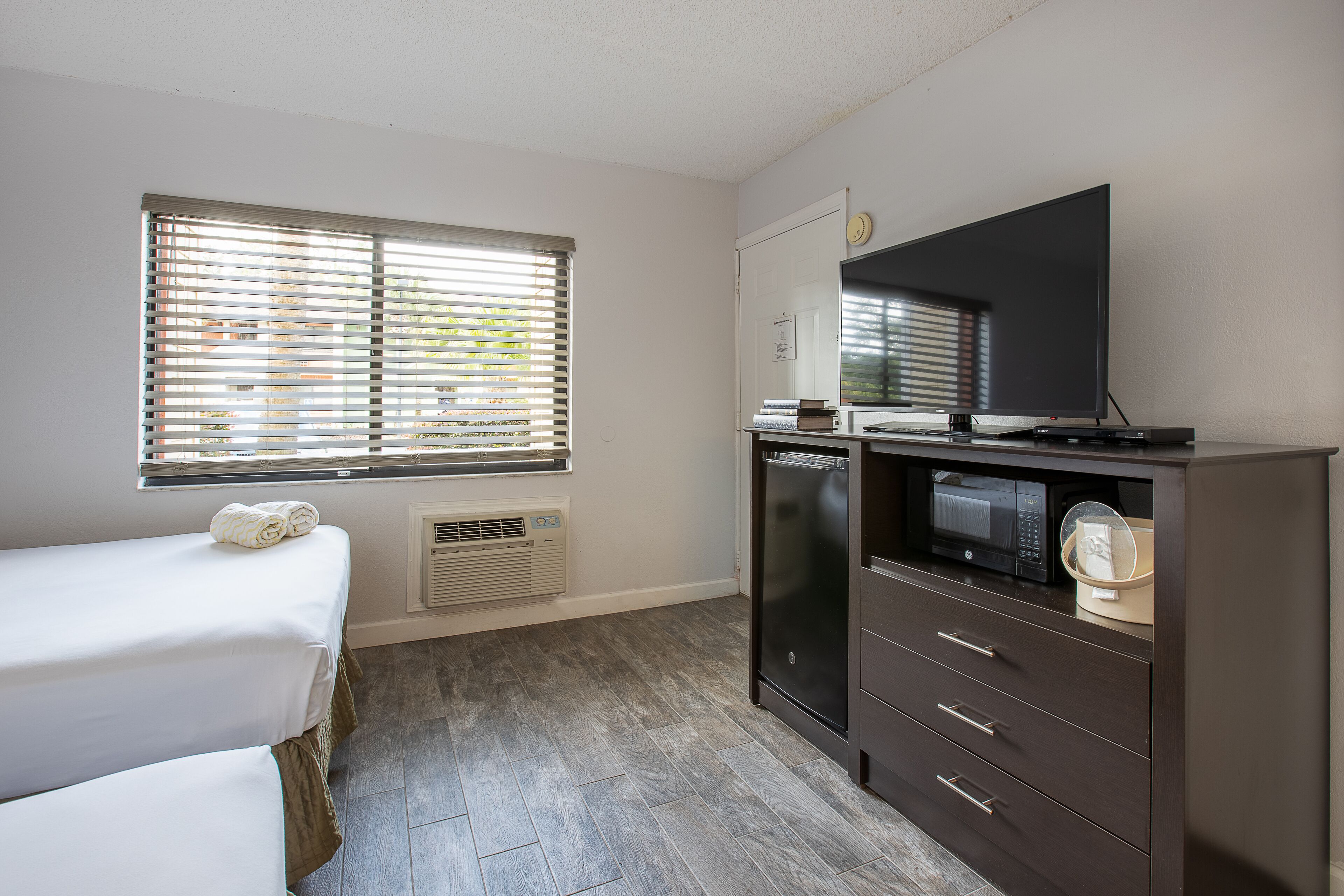 Ein Hotelzimmer mit Bett, Schreibtisch, Flachbildfernseher, Mikrowelle und einem Fenster mit Jalousien.