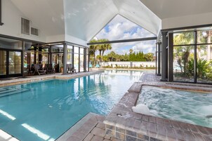 An indoor swimming pool with clear glass walls, a hot tub, and a patio area with lounge chairs.