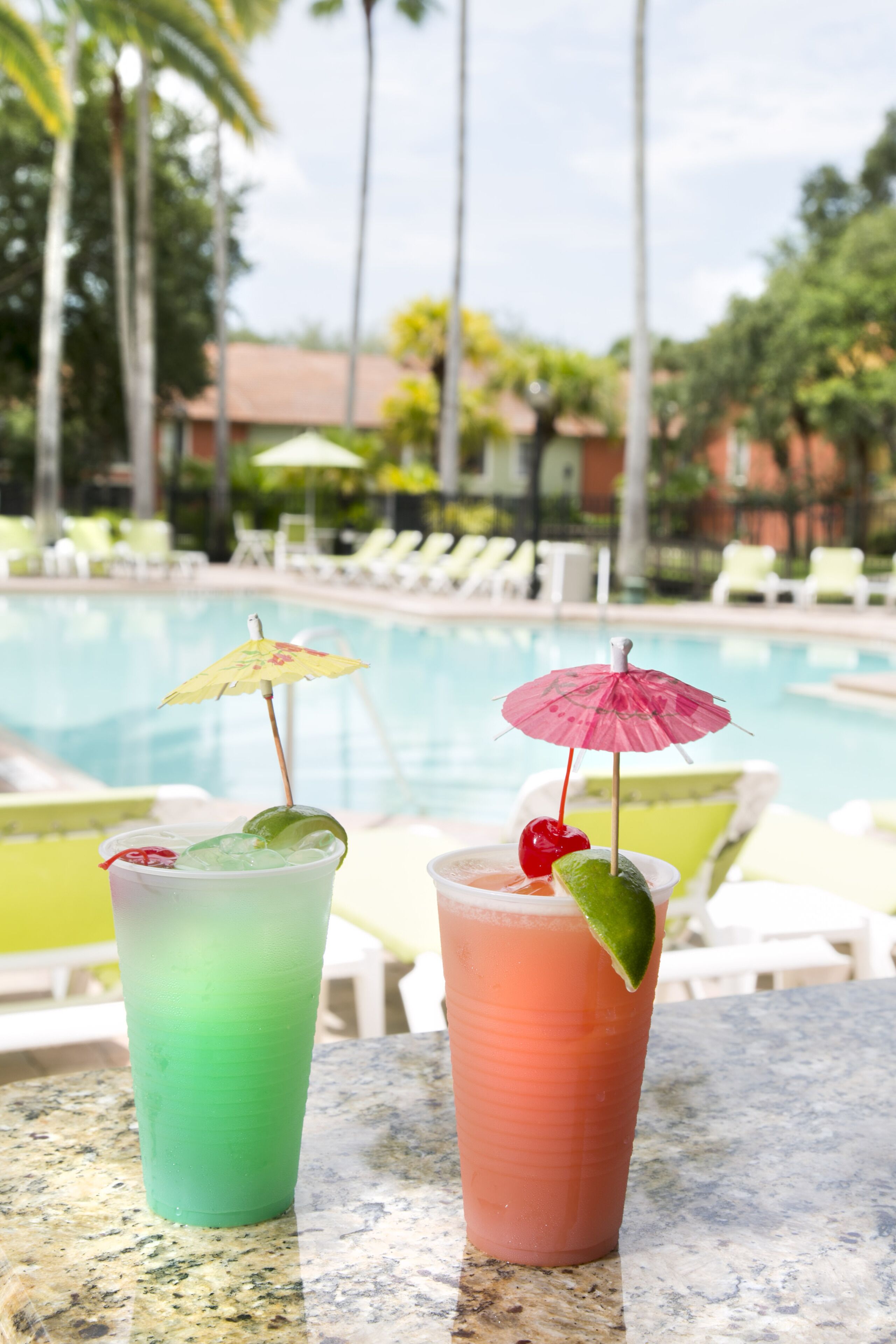 Two drinks with umbrellas and lime slices on a table by a poolside with lounge chairs and palm trees in the background.