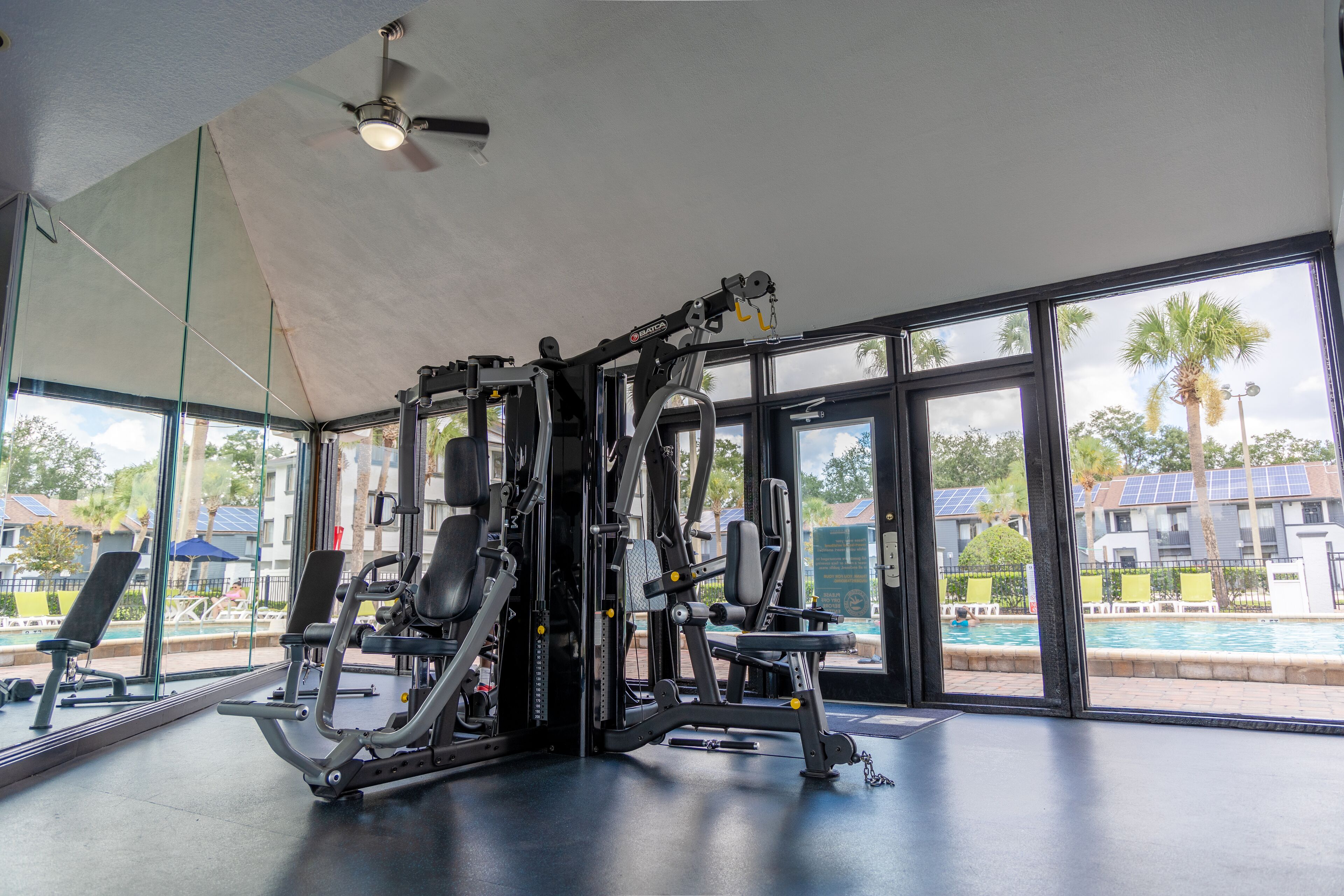 Une salle de sport équipée de matériel d’exercice, dotée de grandes fenêtres et offrant une vue sur une piscine et des bâtiments.