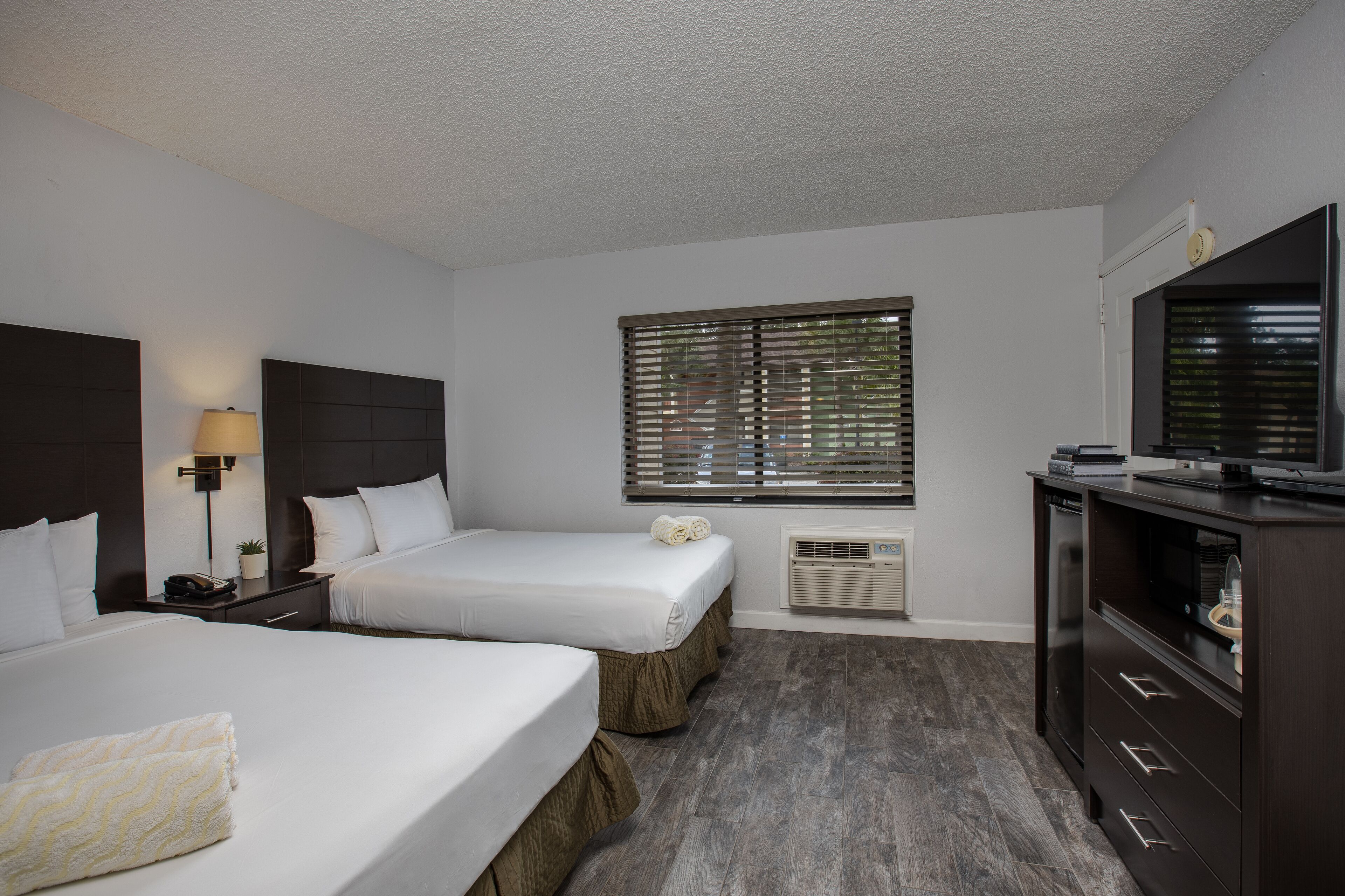 Une chambre d’hôtel avec deux lits, une télévision, un bureau avec une lampe et une fenêtre avec des stores.
