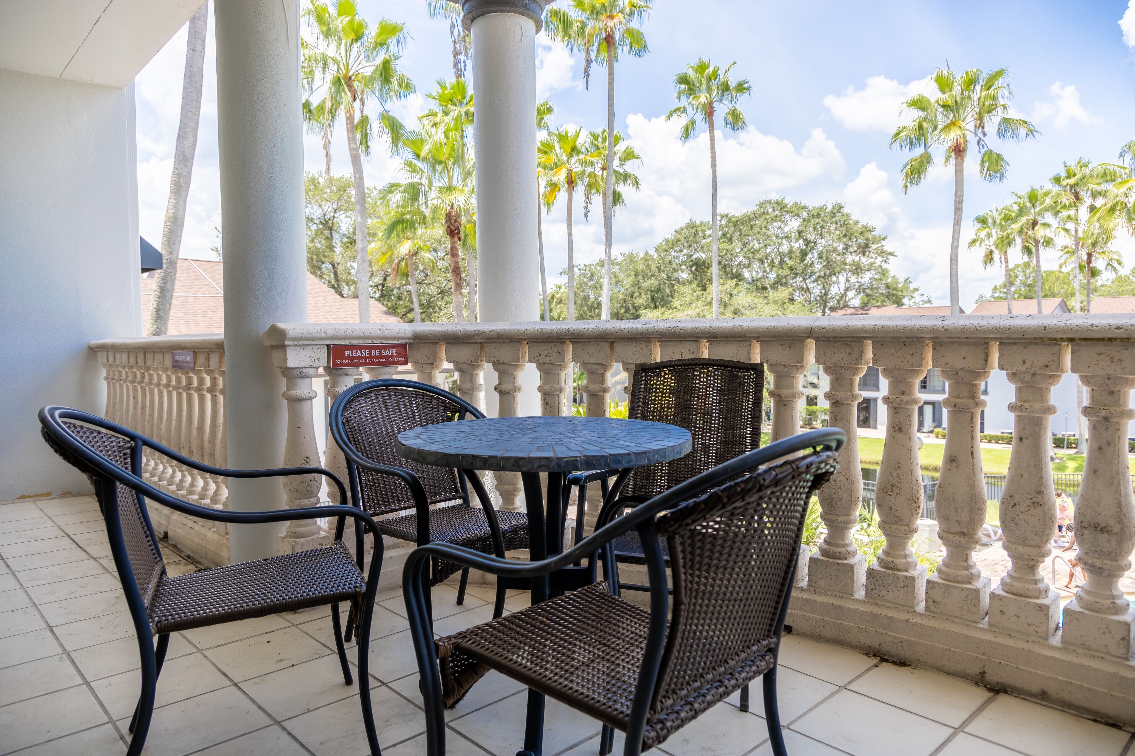 A balcony with a round table and four wicker chairs, overlooking a garden with palm trees and a clear sky.