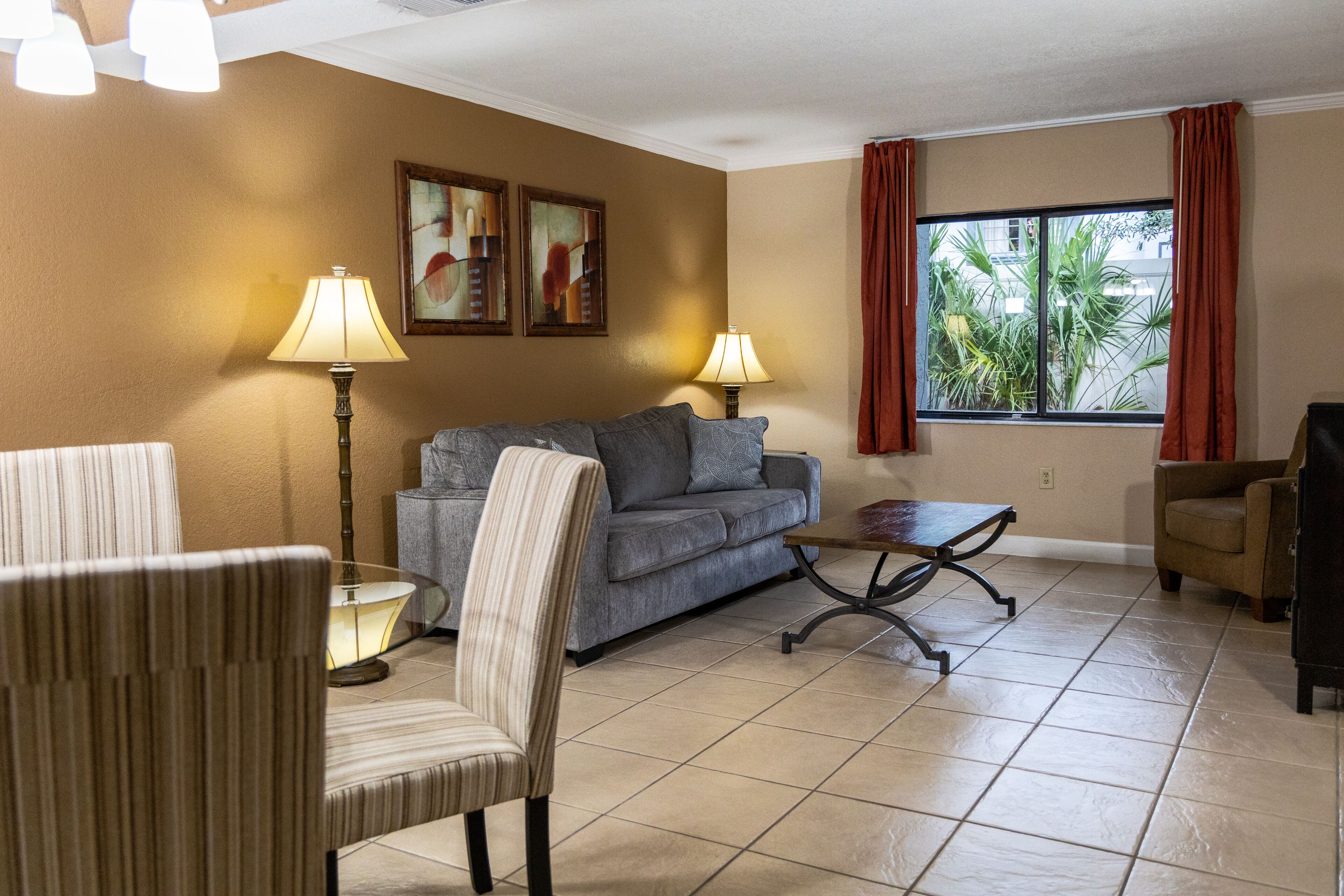 A living room with a sofa, armchairs, a coffee table, and two lamps. There are two paintings on the wall and a window with curtains.
