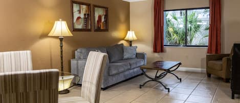 A living room with a sofa, armchairs, a coffee table, and two lamps. There are two paintings on the wall and a window with curtains.