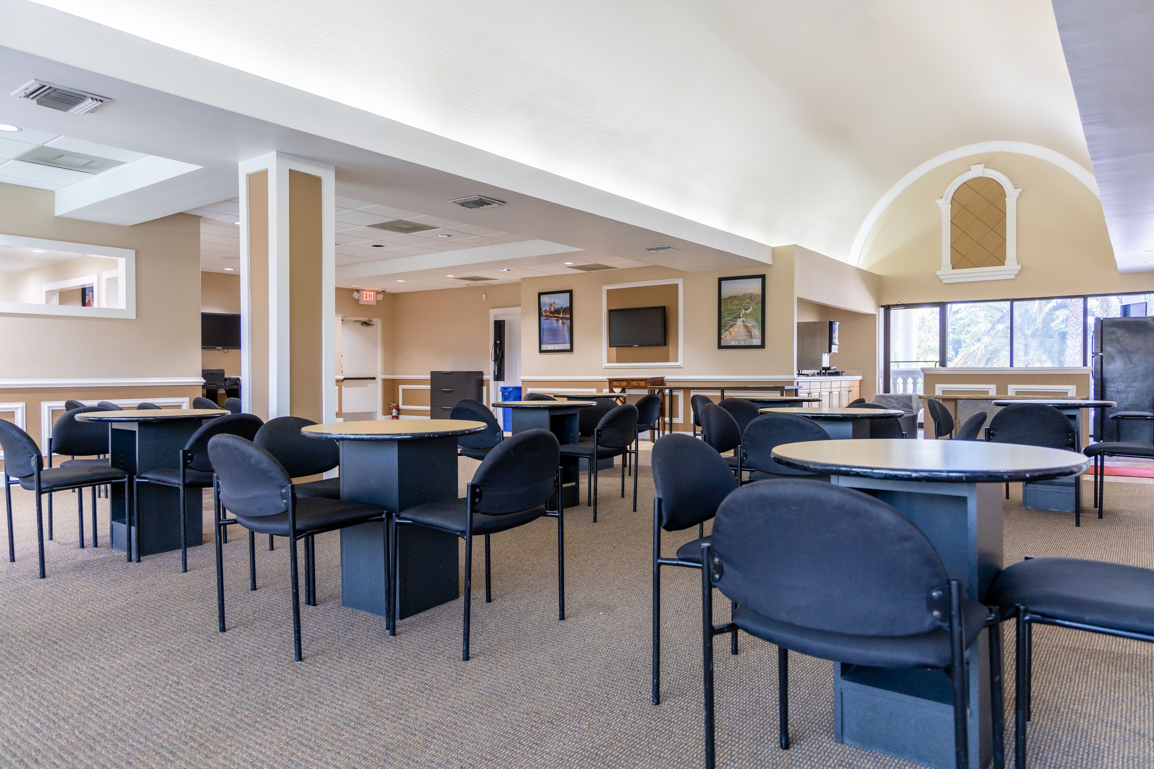 A spacious conference room with multiple tables and chairs, large windows, and a high ceiling with recessed lighting.