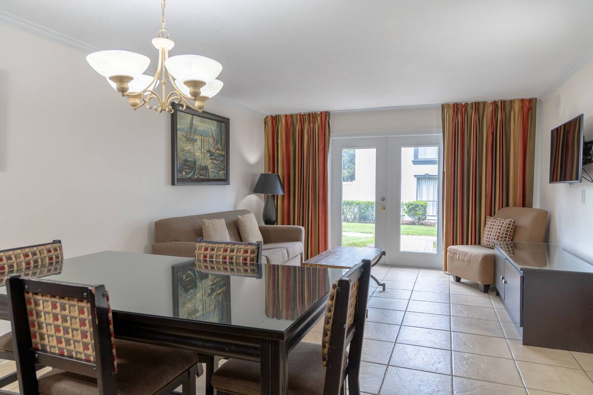 A dining area with a glass table, chairs, a chandelier, a sofa, and a television.