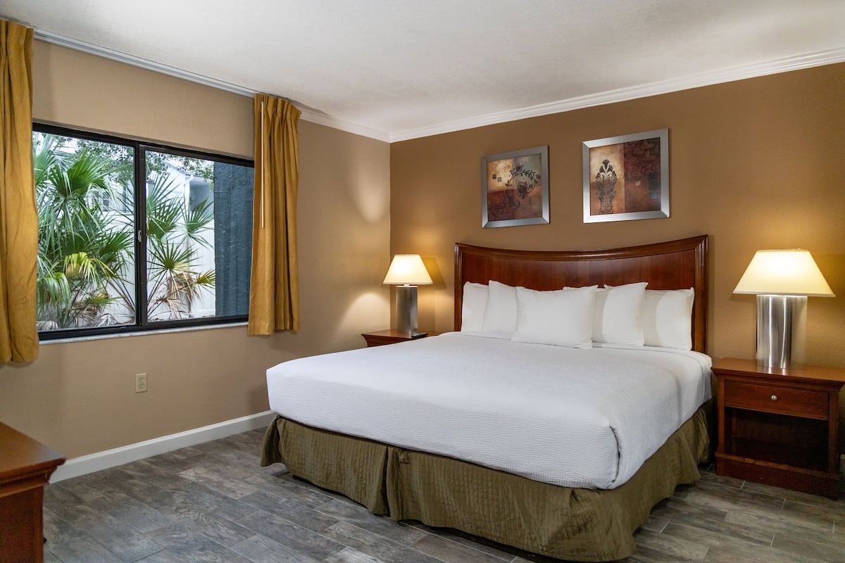 A hotel room with a large bed, two bedside tables with lamps, a window with curtains, and two wall-mounted artworks.