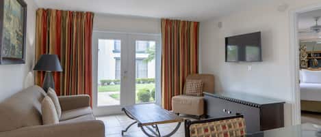 A living room with a sofa, armchair, and a table. There is a television mounted on the wall and a door leading outside.