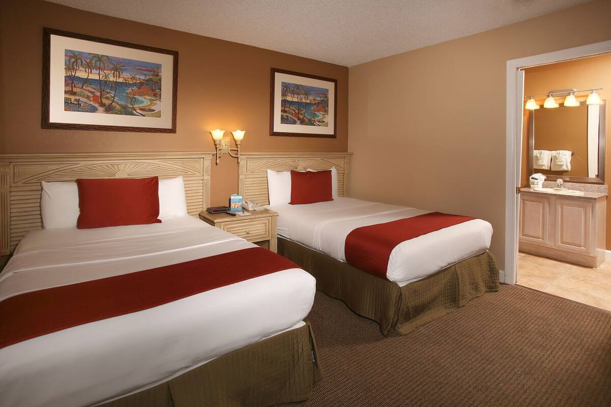 A hotel room with two beds, a nightstand, a lamp, a mirror, and artwork on the walls.