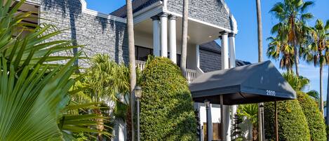 A large, modern building with a stone facade, a covered entrance, and a well-maintained garden with palm trees.