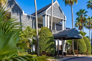 A large, modern building with a stone facade, a covered entrance, and a well-maintained garden with palm trees.
