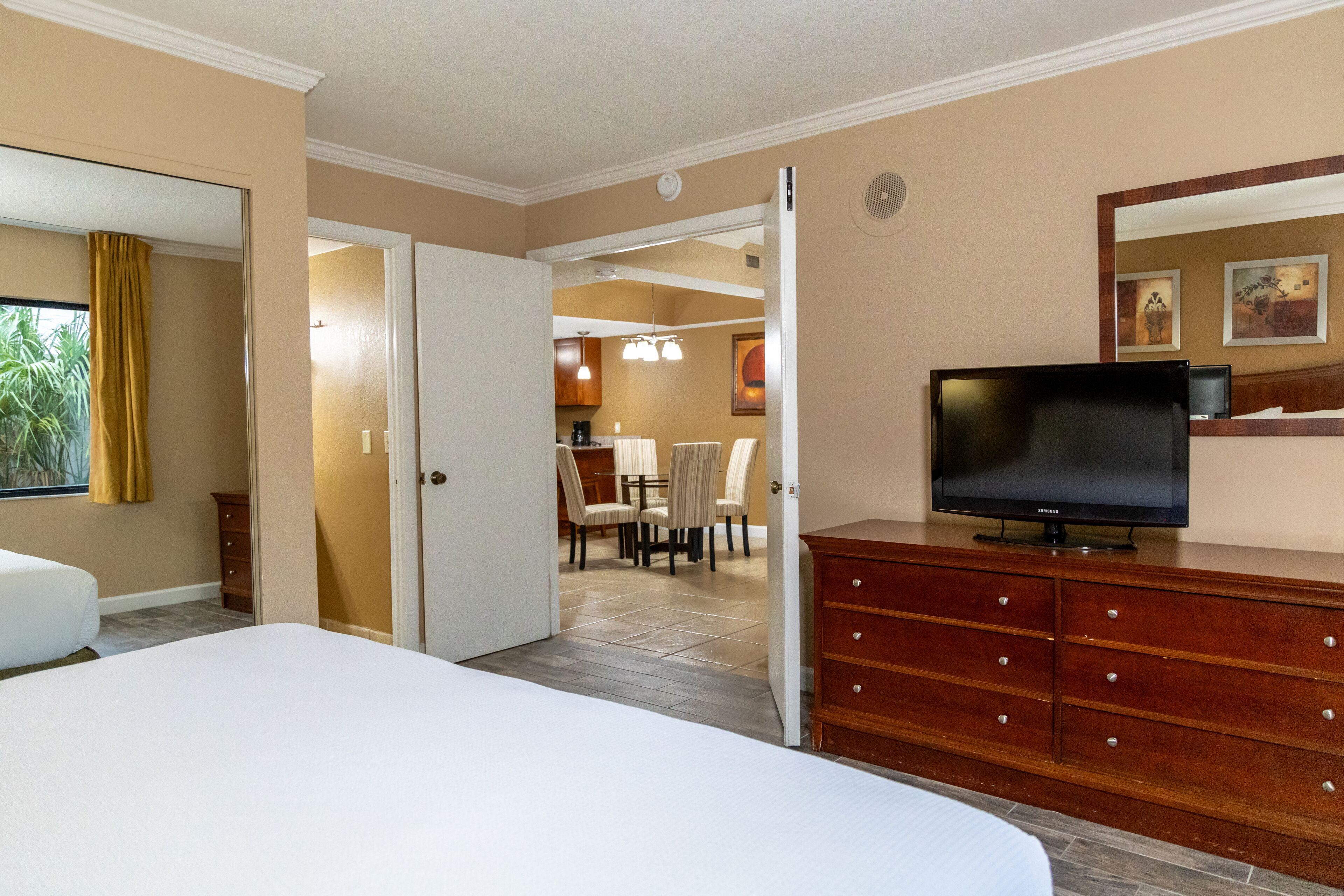 A hotel room with a bed, a TV on a wooden dresser, a dining area with chairs and a table, and a kitchen in the background.