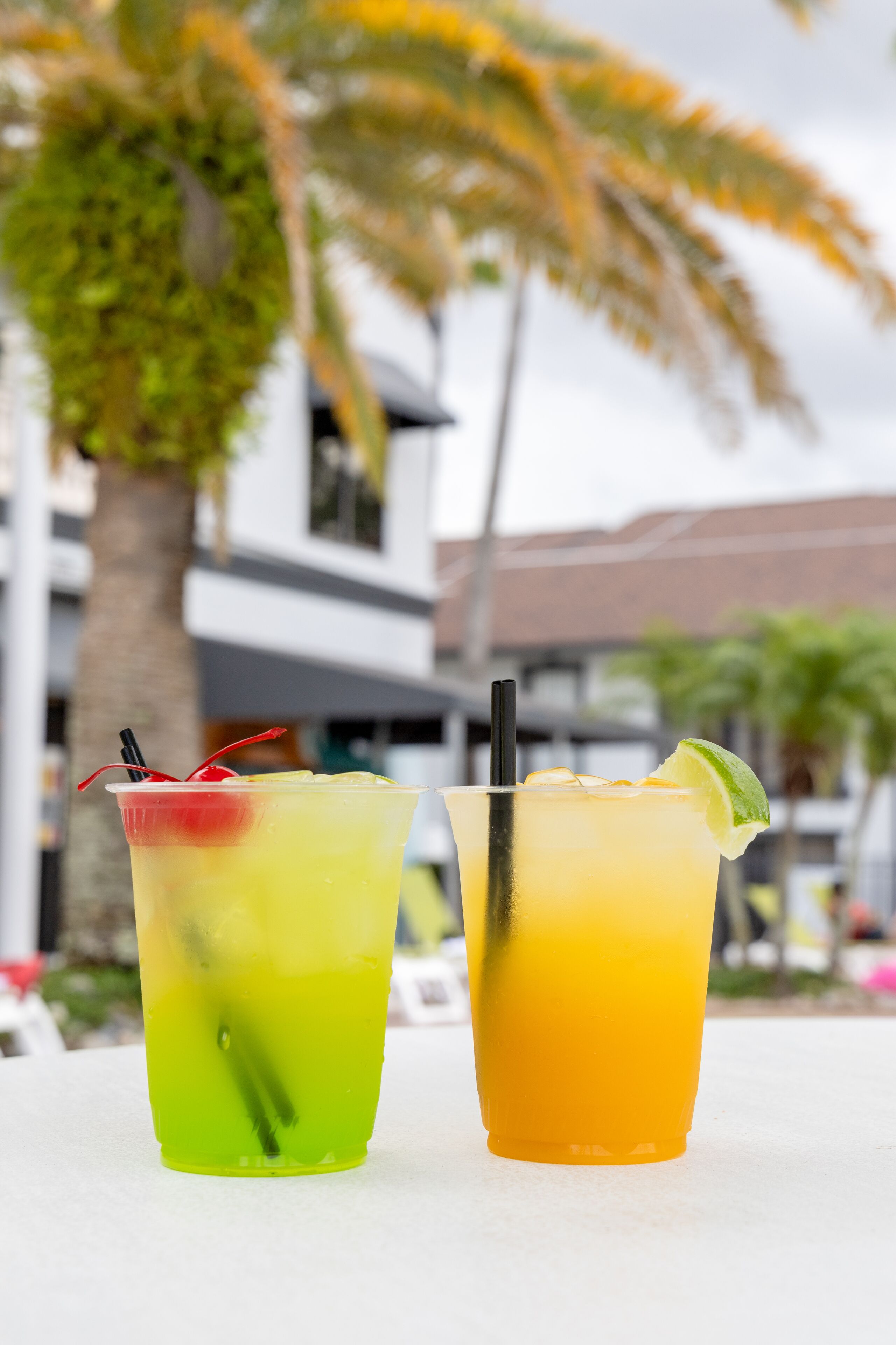 Duas bebidas com canudos e fatias de limão sobre uma mesa, com um fundo desfocado de um edifício e palmeiras.