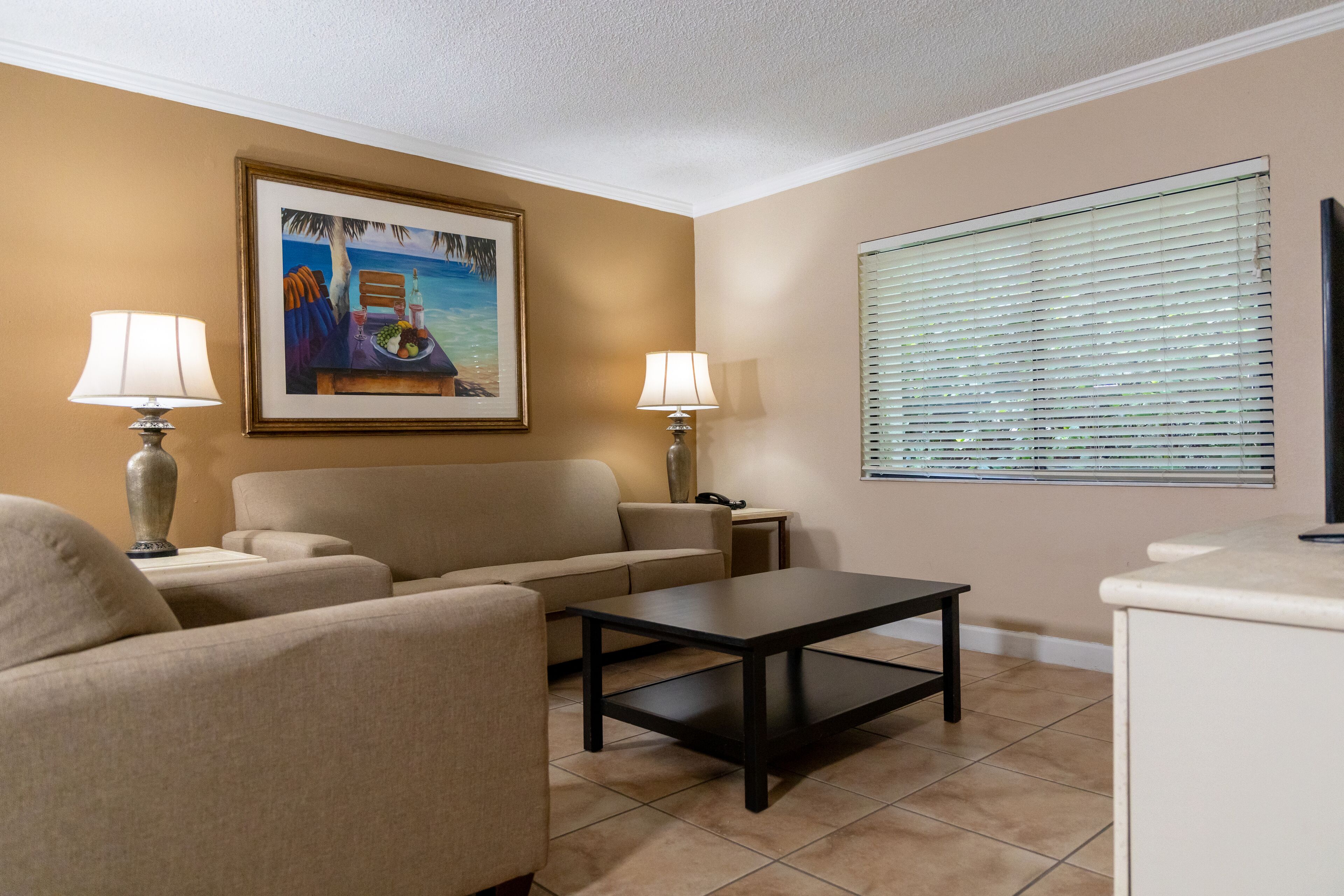 A living room with beige walls, a beige sofa, two lamps, a coffee table, and a painting of a beach scene.